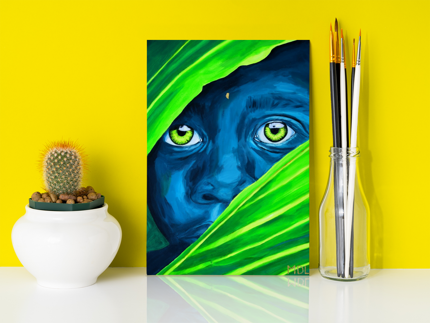 A small colorful art print on canvas of an African Boy, leaning up against a yellow wall on a desk next to some paint brushes and a cactus