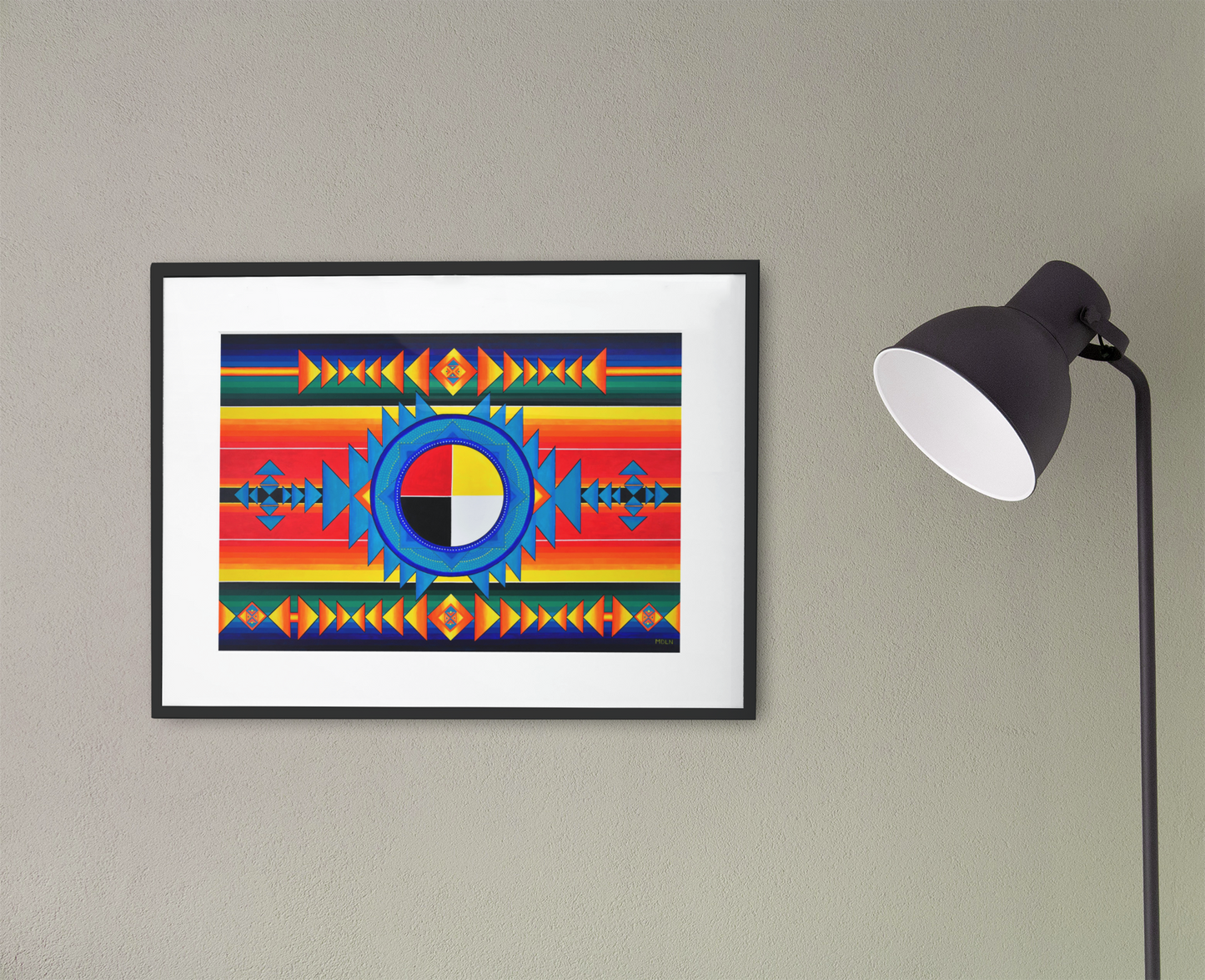 A small but bright Indigenous Medicine Wheel giclee art print, framed in black with white matting, hanging on a wall next to a lamp